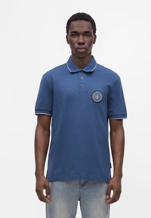 Homme portant un polo bleu à manches courtes avec un emblème sur la poitrine et un jean clair délavé, debout devant un fond blanc uni.