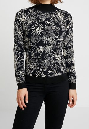 Femme portant un pull noir à manches longues avec des motifs botaniques et d'insectes blancs, associé à un pantalon noir, debout devant un fond uni.