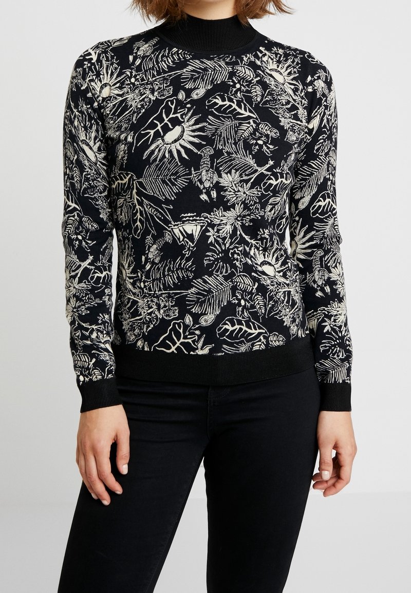 Femme portant un pull noir à manches longues avec des motifs botaniques et d'insectes blancs, associé à un pantalon noir, debout devant un fond uni.
