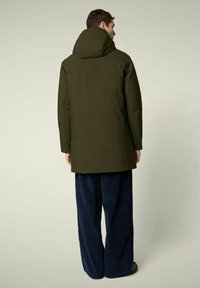 Olijfgroene geïsoleerde parka met een grote capuchon, rechte snit en een ruwe textuur. Gecombineerd met marineblauwe wijde broekspijpen.