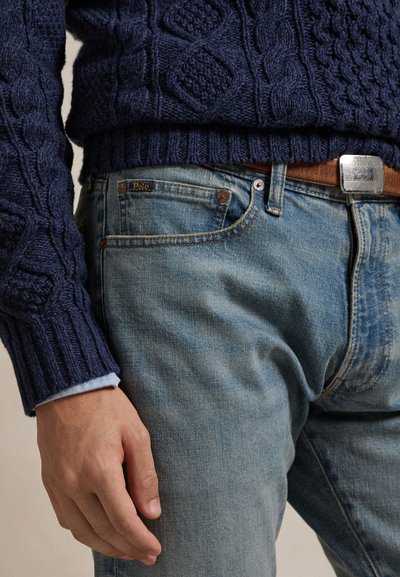 Muž nosící modrý svetr s copánkovým vzorem založený do světle modrých džín Polo s hnědým opaskem, ruka uvolněně podél těla.