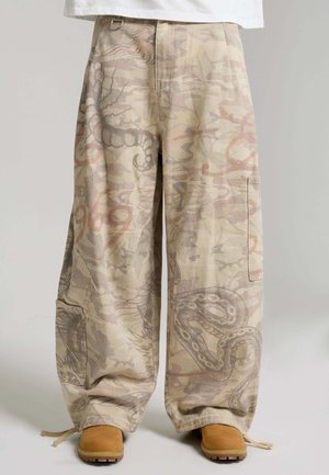 Luźne beżowe spodnie typu cargo z przygaszonymi wzorami węża i abstrakcyjnymi, bocznymi kieszeniami, ściągaczami na dole, noszone z jasnobrązowymi botkami i białym topem.