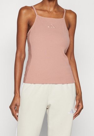 Kobieta ubrana w kurzowo-różowy, prążkowany, bezrękawnik z cienkimi ramiączkami oraz beżowe dresy Adidas z logo.