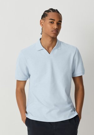 Ung mand med fletninger, iført en lyseblå kortærmet poloshirt og mørke bukser, står med hænderne i lommerne.