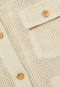 Crèmegekleurde, gestructureerde brei stof met een ruitpatroon, een flapzak en gouden hardware accenten.
