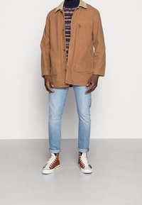 Brun bomullskavaj med en ljusfärgad krage, två stora framfickor, bärs över en mönstrad tröja, i kombination med ljusblå jeans och sneakers.