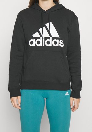 Osoba nosząca czarną bluzę z kapturem Adidas z dużym białym logo oraz niebieskie legginsy Adidas z małym białym logo.