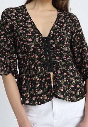 Vrouw draagt een zwarte blouse met roze bloemen, V-hals, knopen aan de voorkant, en witte broek, geknipt ter hoogte van de taille.