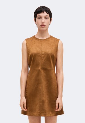 Jeune personne aux cheveux courts et foncés portant une robe marron en daim sans manches, debout devant un fond clair uni.