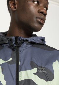 Giacca a vento camouflage in blu navy e verde menta, con zip frontale, cappuccio regolabile e logo ricamato sul petto. Tessuto liscio e leggero.