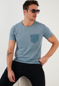 Světle modré tričko s krátkým rukávem, které má texturovanou modrou kapsu, hladkou tkaninu, kulatý výstřih a slim fit. Nošeno s tmavými kalhotami.
