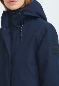 Giacca impermeabile color navy con colletto alto, cappuccio regolabile e patch nera del brand. Presenta dettagli a cordino in contrasto e bottoni a pressione.