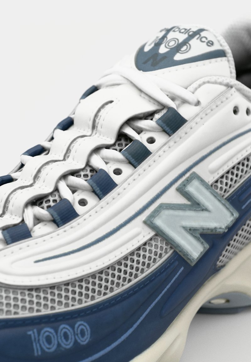 Las zapatillas New Balance 1000 presentan un diseño en blanco y azul, con una parte superior de malla, suela de goma y un prominente logo azul de "N" en el lateral.
