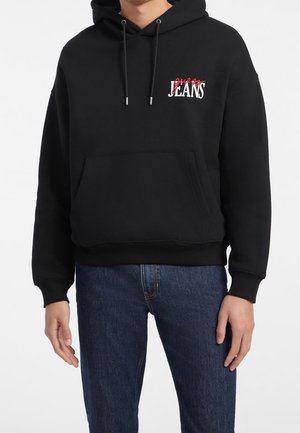 Man draagt een zwarte hoodie met voorvak en een rood-wit "Guess Jeans" logo, gecombineerd met een blauwe spijkerbroek.