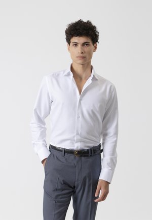 Hombre con cabello rizado, vestido con una camisa blanca de vestir y pantalones grises con un cinturón negro, de pie frente a un fondo claro y liso.