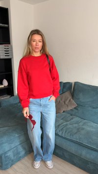 Červená mikina, svetlomodré rifle, držiace červené slnečné okuliare. Pozadie tvorí tyrkysová pohovka s rebrovanou textúrou a police. Neformálne oblečenie.