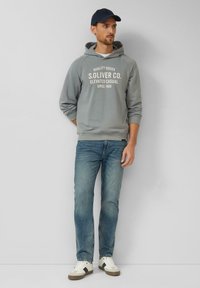 Grauer Kapuzenpullover mit weißem Textlogo, kombiniert mit hellblauen Jeans und weißen Sneakers mit grauen Akzenten. Schlichtes Design, lässige Passform.
