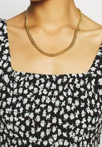 Collar de cadena de oro con eslabones entrelazados, con un acabado suave y pulido. Llevado con un vestido negro de estampado floral.