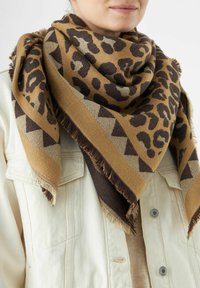 Lepardschildering sjaal in bruin en beige met geometrische accenten, met franjes aan de randen, over een lichtgekleurde, knopenjasje gedrapeerd.