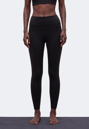 Vrouw staat blootsvoets op een yogamat, draagt zwarte leggings met hoge taille en een zwart crop top tegen een effen achtergrond.