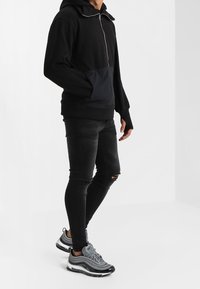 Felpa con zip nera e tasca blu a contrasto, abbinata a jeans skinny neri strappati e scarpe da ginnastica grigie con superficie testurizzata.