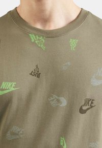 Camiseta color caqui con varios logos y frases de Nike impresos en verde y gris. Diseño de cuello redondo con una tela suave y texturizada.