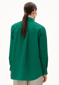 Grüne Bluse mit einem Rückenschlitz, langen Ärmeln, Knopfmanschetten und einem leicht geschwungenen Saum. Hergestellt aus glattem Stoff.