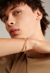 Un bracelet en métal doré sur un poignet, avec une surface lisse et brillante. La personne porte une peau claire et est vêtue d'une chemise beige.