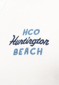 Broderie sur tissu blanc avec le texte « HCO Huntington Beach » en fil bleu et marine, disposé en trois lignes centrées.