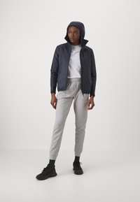 Marineblå hættepufferjakke, let stof, grå sweatpants med manchetter, sorte sneakers, stående mod en ensfarvet hvid baggrund.