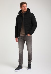 Sort pufferjakke med hætte og frontlynlås, parret med en beige ribbet sweater og grå jeans. Fodtøjet er mørke overknæ sneakers.
