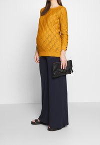 Senapsgul stickad tröja med diamants mönster, parat med marinblå vida byxor, håller en svart texturerad väska. Svarta sandaler.