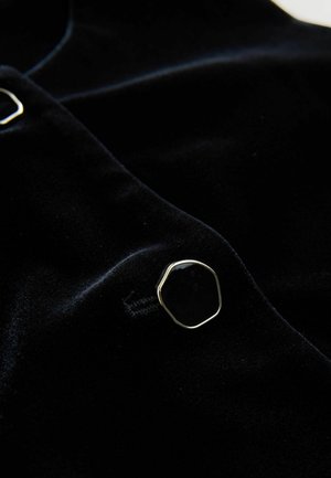 Černá sametová tkanina s jemnou texturou, která obsahuje zlatě tónovaný, osmiúhlý detail knoflíku. Na materiálu je patrný jemný lesk.