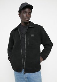 Black fleece jacket with a zippered front, high collar, and chest pocket. Features soft texture and a fitted design, paired with light blue jeans.
