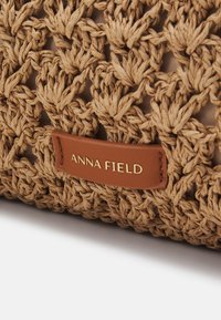 Tkaná slámová taška v přírodní béžové barvě s texturovaným vzorem. Obsahuje hnědou koženou visačku s vyraženým nápisem "ANNA FIELD" zlatou barvou.