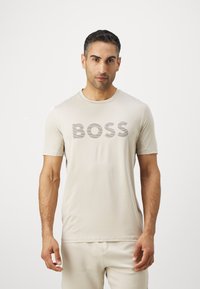 Camiseta de manga corta color beige con un destacado logo "BOSS" en gris oscuro. Textura suave y sencillo escote redondo.
