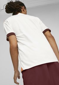 Valkoinen urheilupaita, jossa on maroonit yksityiskohdat hihoissa, yhdistetty tummamarooniin shortsiin. Paidassa on teksturoitu pinta ja sivussa oranssi yksityiskohta.