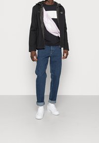 Giacca nera con cappuccio sopra un maglione blu navy con una striscia bianca, abbinata a jeans blu arrotolati e sneaker bianche. Presenta una borsa a tracolla rosa.