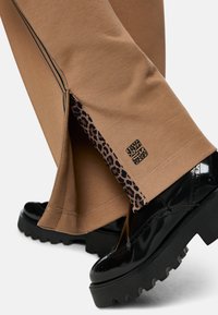 Tan sweatpants with leopard print side slits and black stripe detail. Features a black logo and paired with shiny black platform shoes.