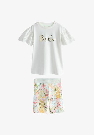 Hvid bluse med korte ærmer og blonder, med guldfarvet tekst "Baker by Ted Baker" kombineret med blomstrede pastelfarvede shorts.