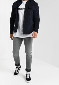 Námořnická košile na knoflíky s dvěma náprsními kapsami, přepnutá přes bílé tričko s potiskem. Šedé džíny a černé tenisky dotvářejí celkový vzhled.