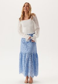 Vit spets långärmad topp kombinerad med en blå spets maxikjol med utsvängd nederkant och blommönster; modellen bär beige sandaler.