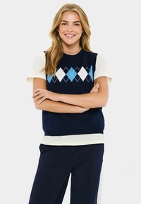 Hymyilevä nainen, jolla on vaaleat hiukset, pukee navy argyle-sweaterin valkoisen lyhythihaiseksi paidan ja navy-housujen päälle, seisten kädet ristissä.
