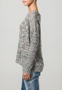 Maglione lavorato a maglia grigio e bianco con una vestibilità morbida, maniche a coste e un motivo testurizzato, mostrato di lato con jeans blu casual.