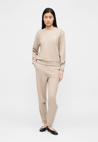 Μπεζ φούτερ και αντίστοιχο παντελόνι φόρμας από απαλό ύφασμα, με λογότυπο στο στήθος και διακοσμητικά σχέδια στα πλάγια. Λείο υλικό.
