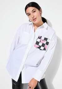 Camicia bianca con bottoni, dotata di una tasca a scacchi con cuori rosa. Presenta maniche lunghe e una vestibilità ampia, realizzata in tessuto leggero.