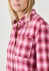 Różowa koszula w kratę z guzikami i kołnierzykiem, wyposażona w kieszeń na piersi, o fakturowanej tkaninie i różnych odcieniach różowego i białego w wzorze.