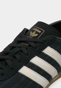 Μαύρο αθλητικό παπούτσι με λευκές ρίγες, διαθέτει υφή στην επάνω επιφάνεια, χρυσό λογότυπο και επίπεδο σύστημα κορδονιών. Η σόλα έχει ανοιχτό καφέ χρώμα.