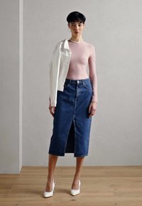 Hellrosa strukturierte Langarmbluse, hochgeschnittener blauer Jeansrock mit seitlichem Schlitz, weiße Jeansjacke über die Schulter geworfen, weiße High Heels.