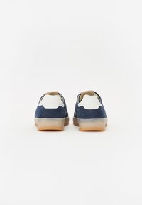 Marineblå ruskinnsneakers med hvite og brune detaljer, gjennomsiktige gummisåler og sydd detaljer ved hælen. Rund form med lav profil.